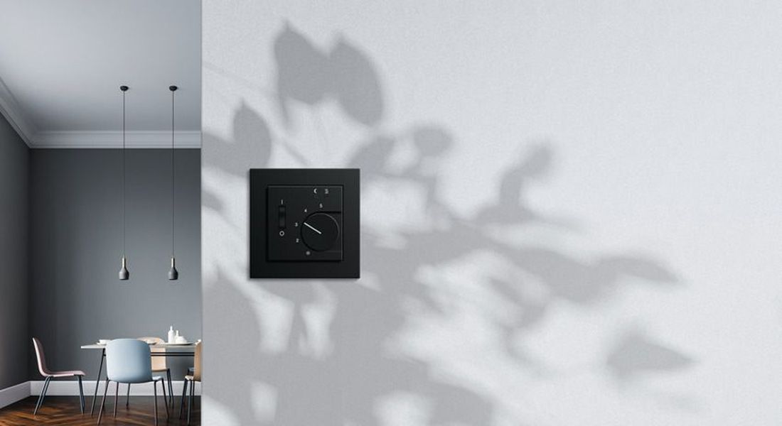 Gira Raumthermostat E2 in Schwarz matt an weißer Wand, Schatten von Blättern, moderner Essbereich im Hintergrund.